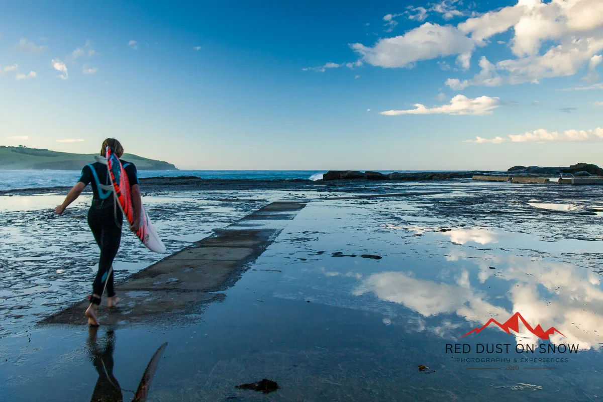 Surfer carrying her board across the rock platform at Werri Beach, Gerringong, with clouds reflected in the pools